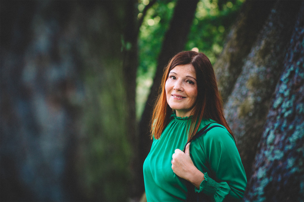Kansanedustaja Sari Sarkomaa, taustalla vihreää ja puunrunkoja.